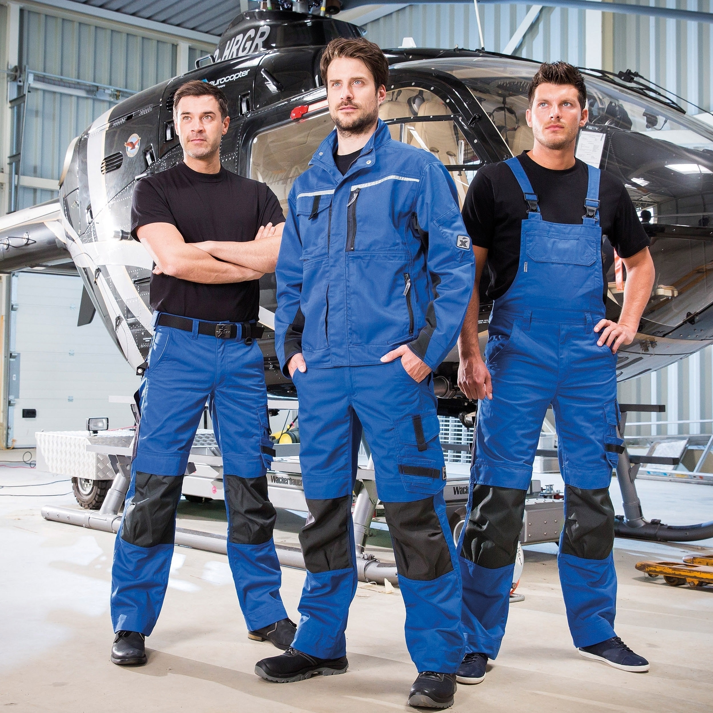 Drei Männer tragen moderne Arbeitskleidung in Blau und Schwarz mit reflektierenden Details und posieren in einer Halle vor einem Hubschrauber.