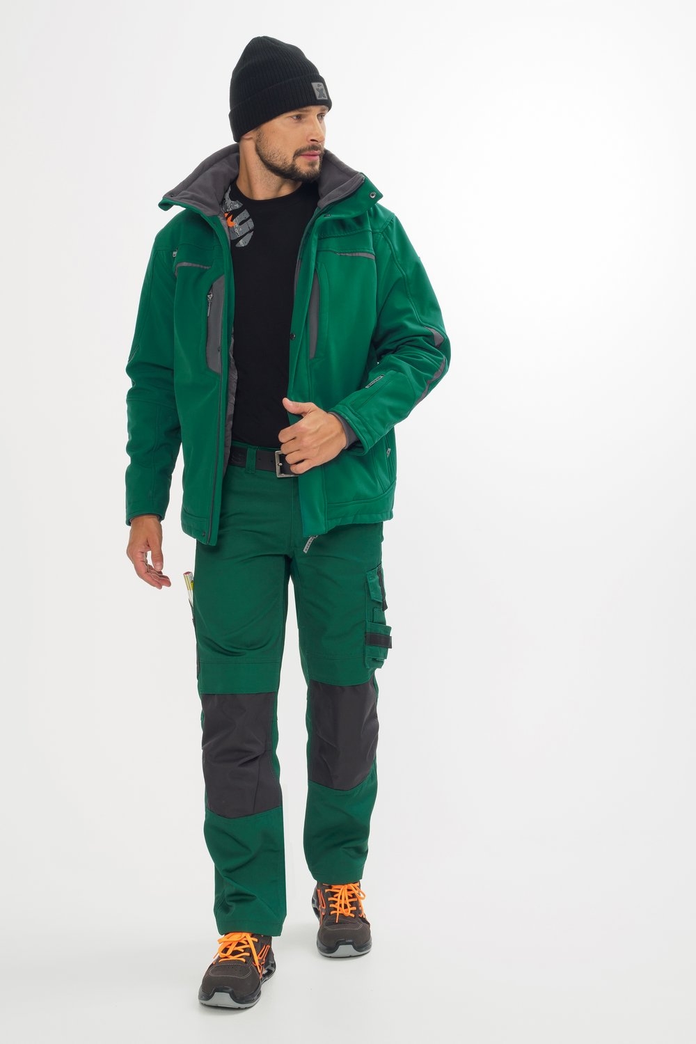 Mann trägt grüne Arbeitsjacke und Hose mit grauen Knieverstärkungen, schwarze Mütze und orange-schwarze Sicherheitsschuhe.