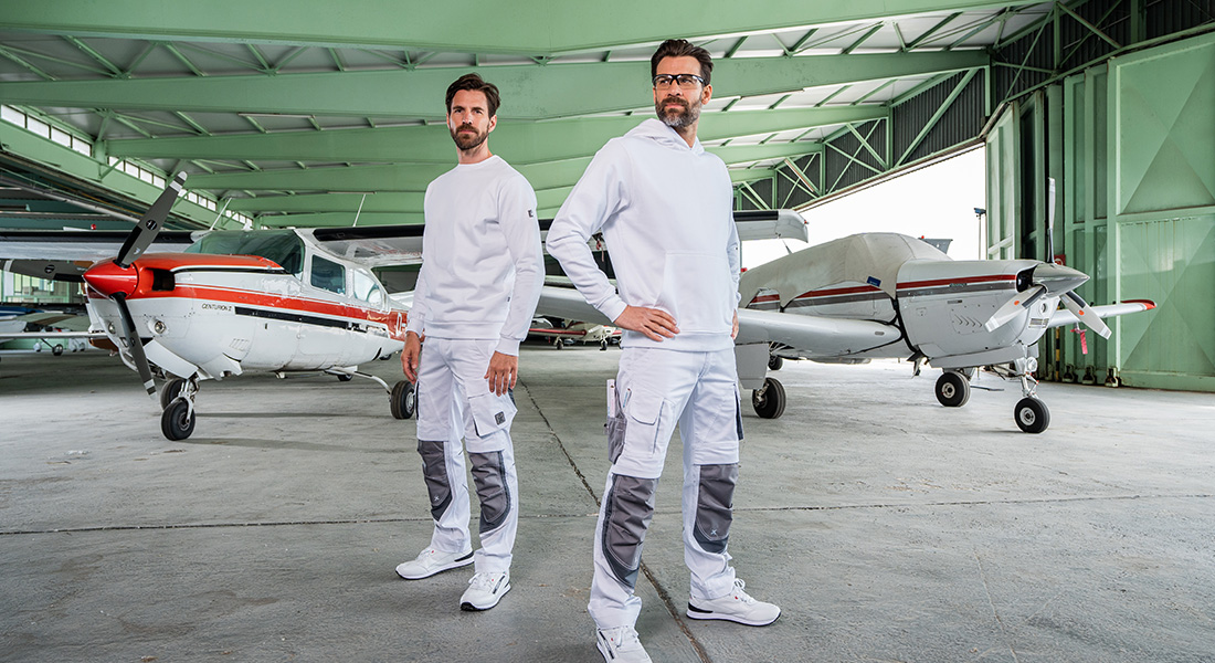 Zwei Männer in weiß-grauer Arbeitskleidung stehen selbstbewusst in einem Flugzeughangar zwischen zwei kleinen Propellermaschinen.