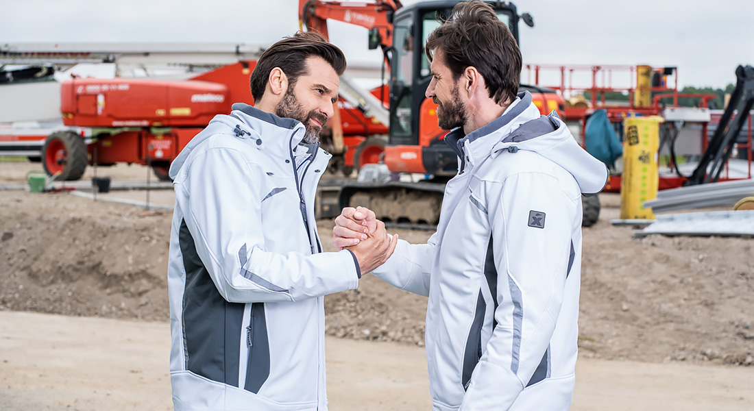 Zwei Männer auf einer Baustelle geben sich die Hand. Beide tragen weiße und graue Arbeitsjacken. Im Hintergrund sind Baumaschinen und Baumaterialien zu sehen.
