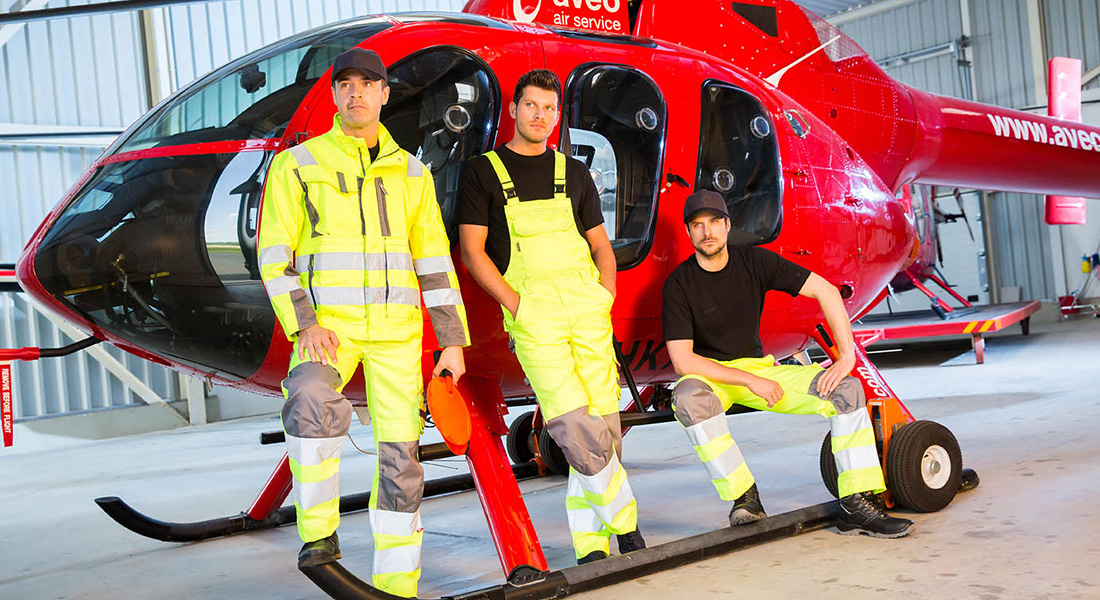 Drei Männer in gelber Warnschutzkleidung posieren vor einem roten Hubschrauber in einer Werkhalle.