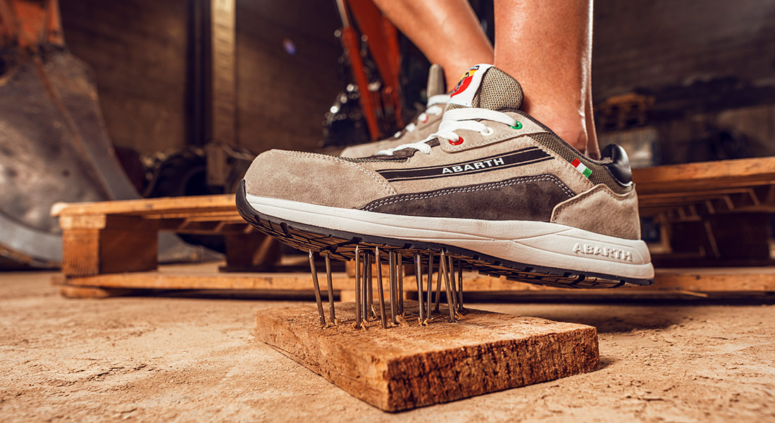 Grauer Abarth-Arbeitsschuh steht auf einem Brett mit herausragenden Nägeln und demonstriert seine durchtrittsichere Sohle in einer Werkstattumgebung.