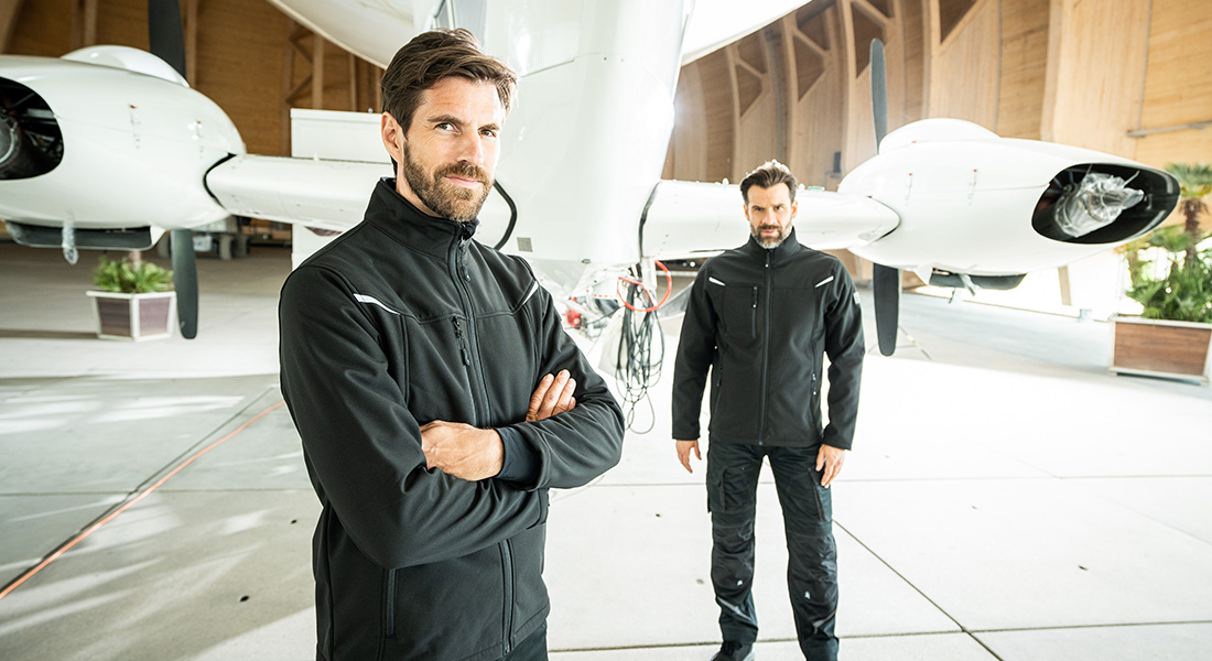 Zwei Männer in schwarzer Arbeitskleidung stehen in einem Hangar vor einem weißen Flugzeug. Einer steht mit verschränkten Armen im Vordergrund, der andere etwas weiter hinten.