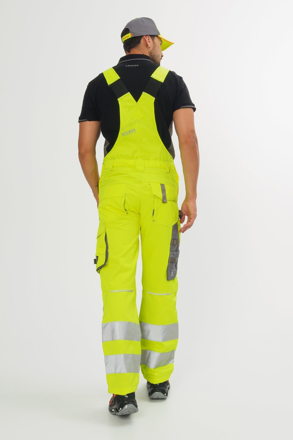 Rückansicht Mann mit gelber Latzhose mit silbernen Reflexstreifen und grauen Einsätzen und schwarzem Poloshirt.
