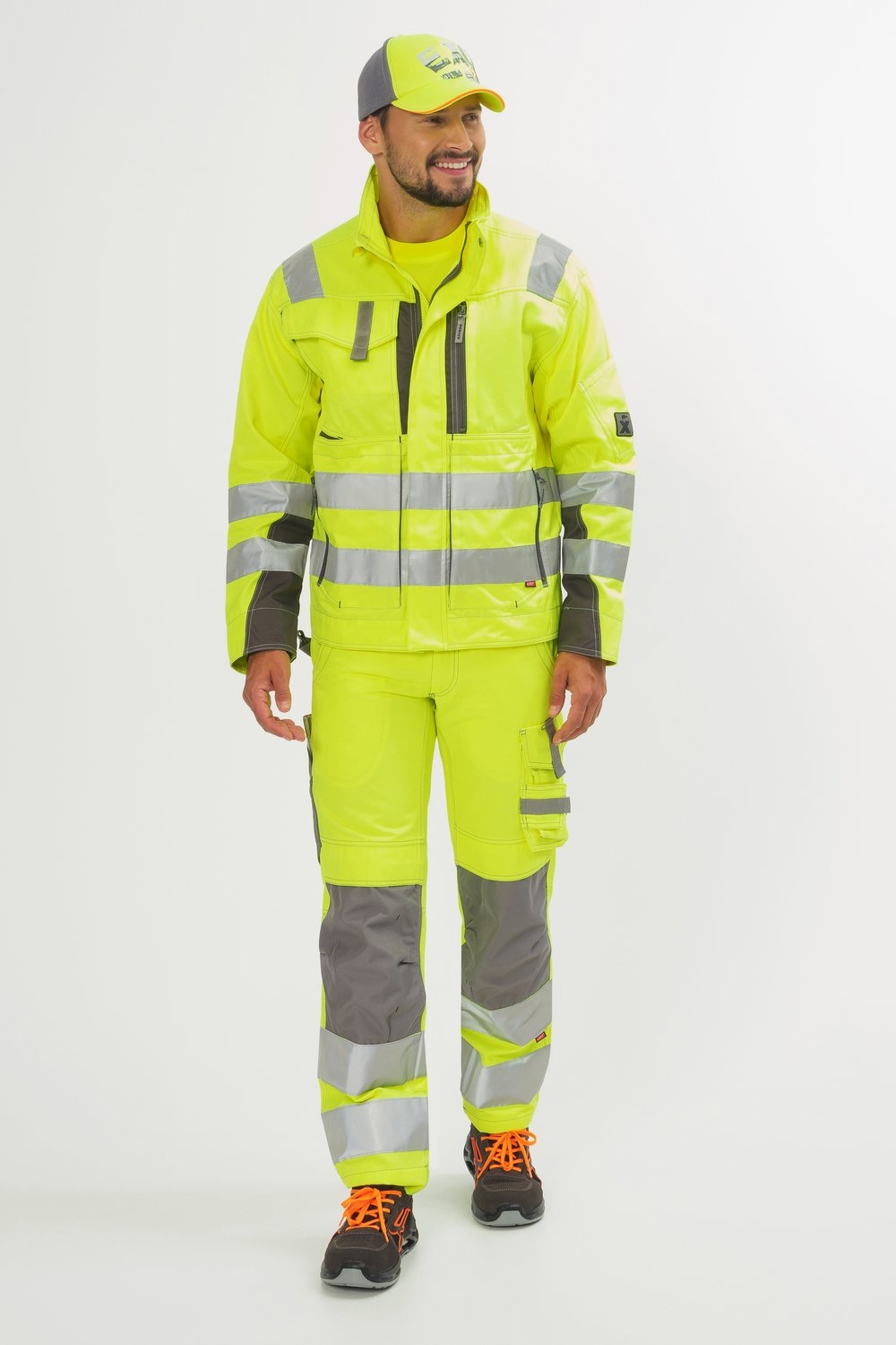 Mann trägt gelbe Arbeitsjacke und Hose mit silbernen Reflexstreifen, grauen Knieeinsätzen, Kappe und Sicherheitsschuhen.