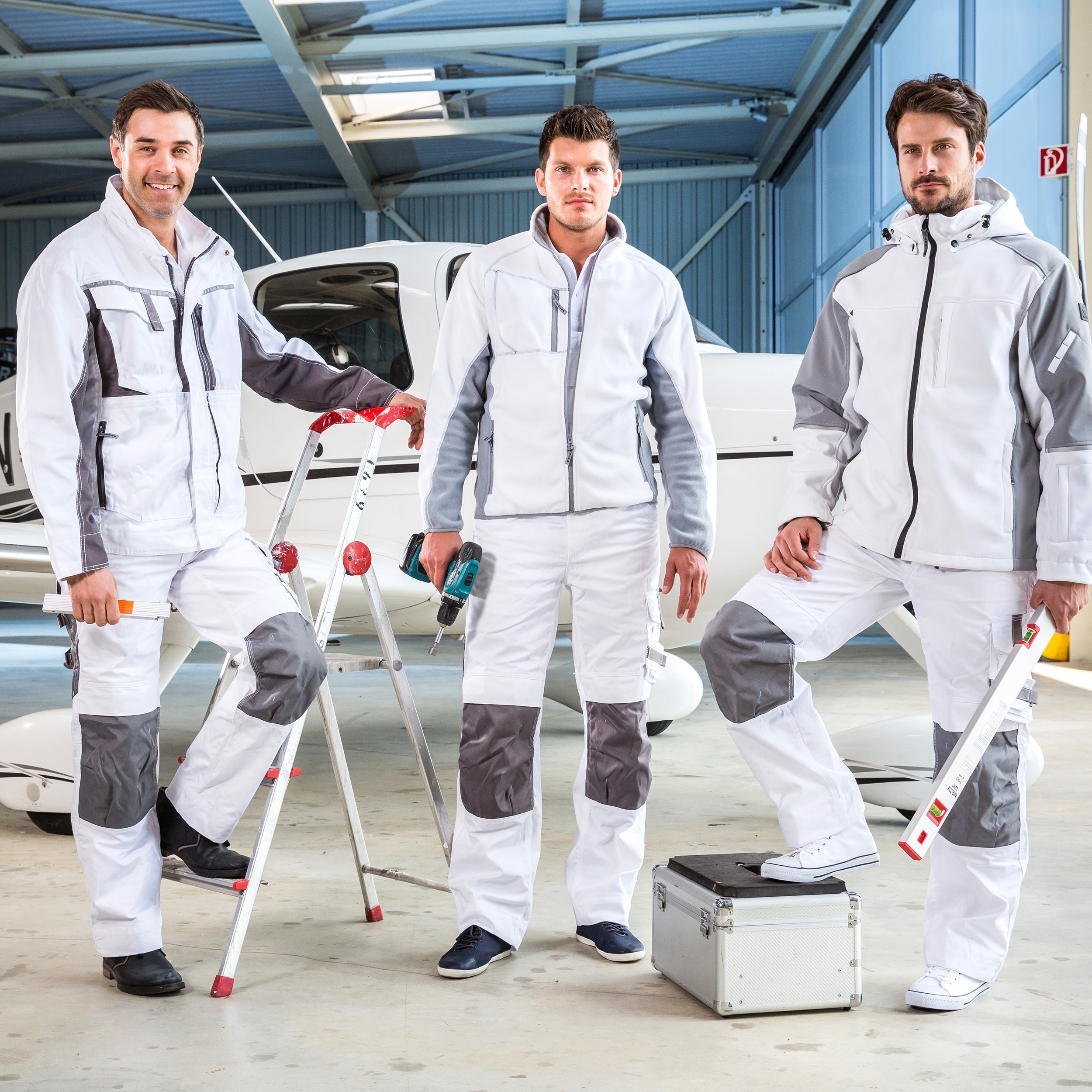 Weiße Arbeitskleidung mit grauen Einsätzen – drei Handwerker mit Werkzeug in Werkstatt vor Flugzeug, EXCESS Workwear.