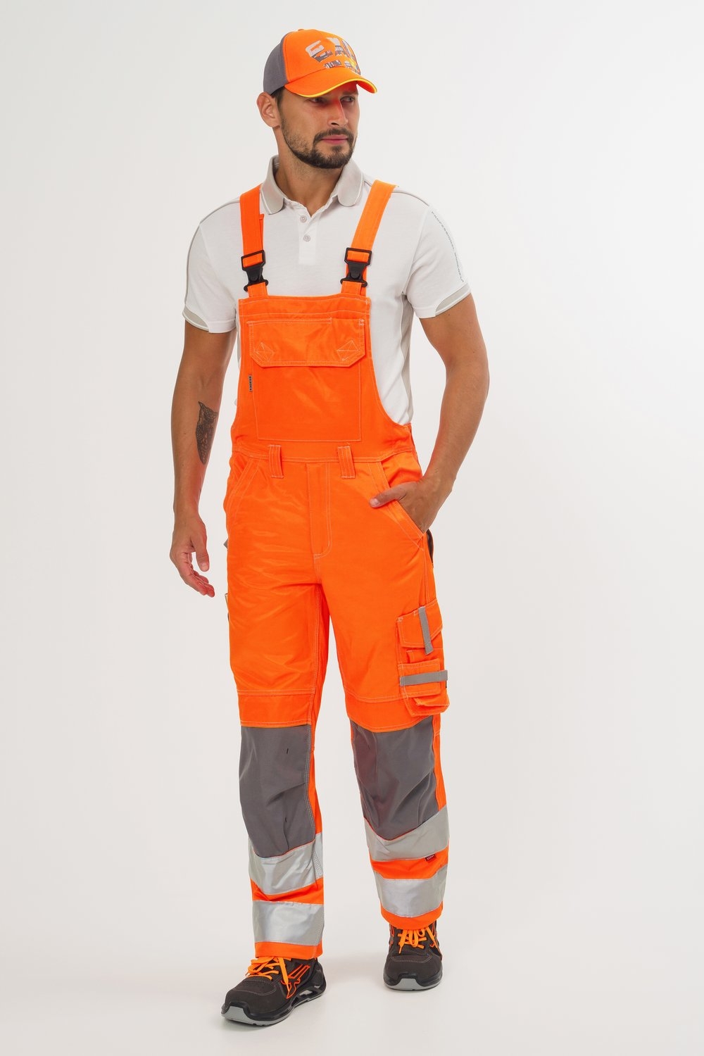 Mann in orangefarbener Latzhose mit Reflexstreifen und weißem Poloshirt und orangefarbener Excess-Cap auf dem Kopf.