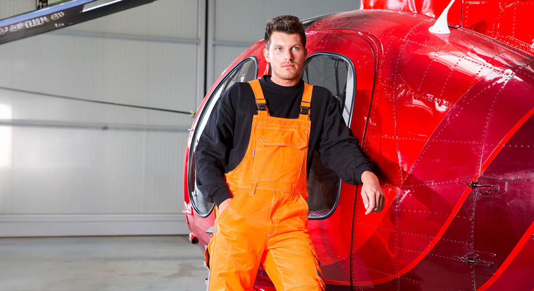 Mann in orangefarbener Arbeitslatzhose und schwarzem Pullover lehnt an einem roten Hubschrauber in einer Werkhalle.