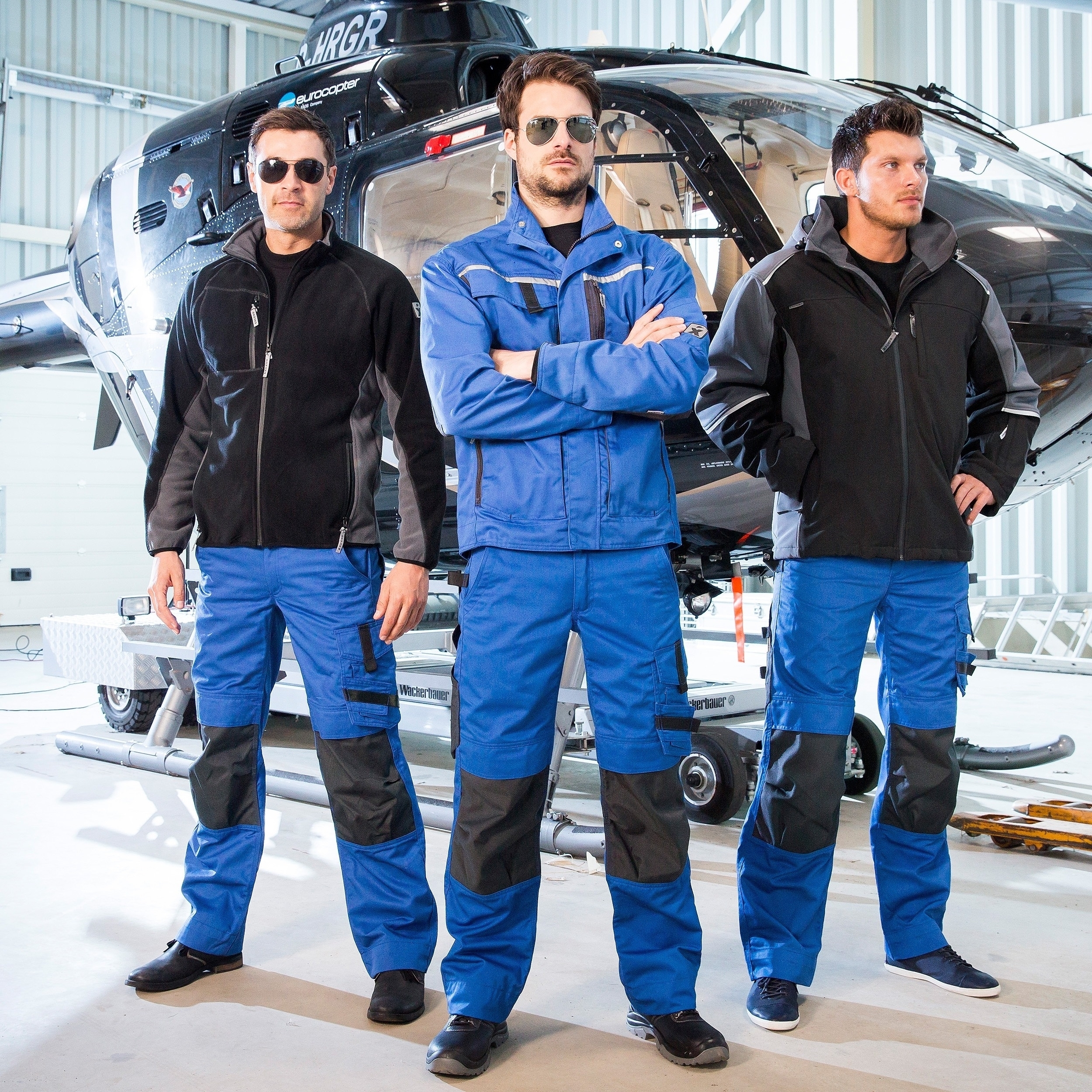 Blaue und schwarze Arbeitskleidung – robuste Berufsmode für Handwerk & Technik, präsentiert von drei Männern vor Hubschrauber.