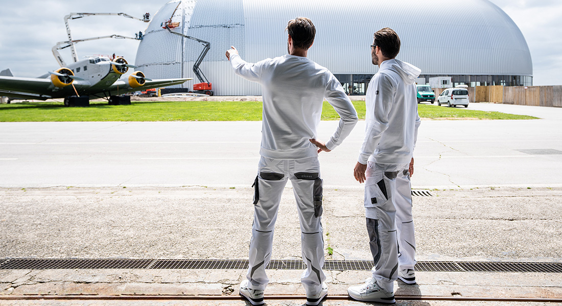 Zwei Männer in weiß-grauer Arbeitskleidung stehen mit dem Rücken zur Kamera und blicken auf ein großes Hangargebäude, während einer von ihnen in die Ferne zeigt. Links ist ein historisches Flugzeug zu sehen.