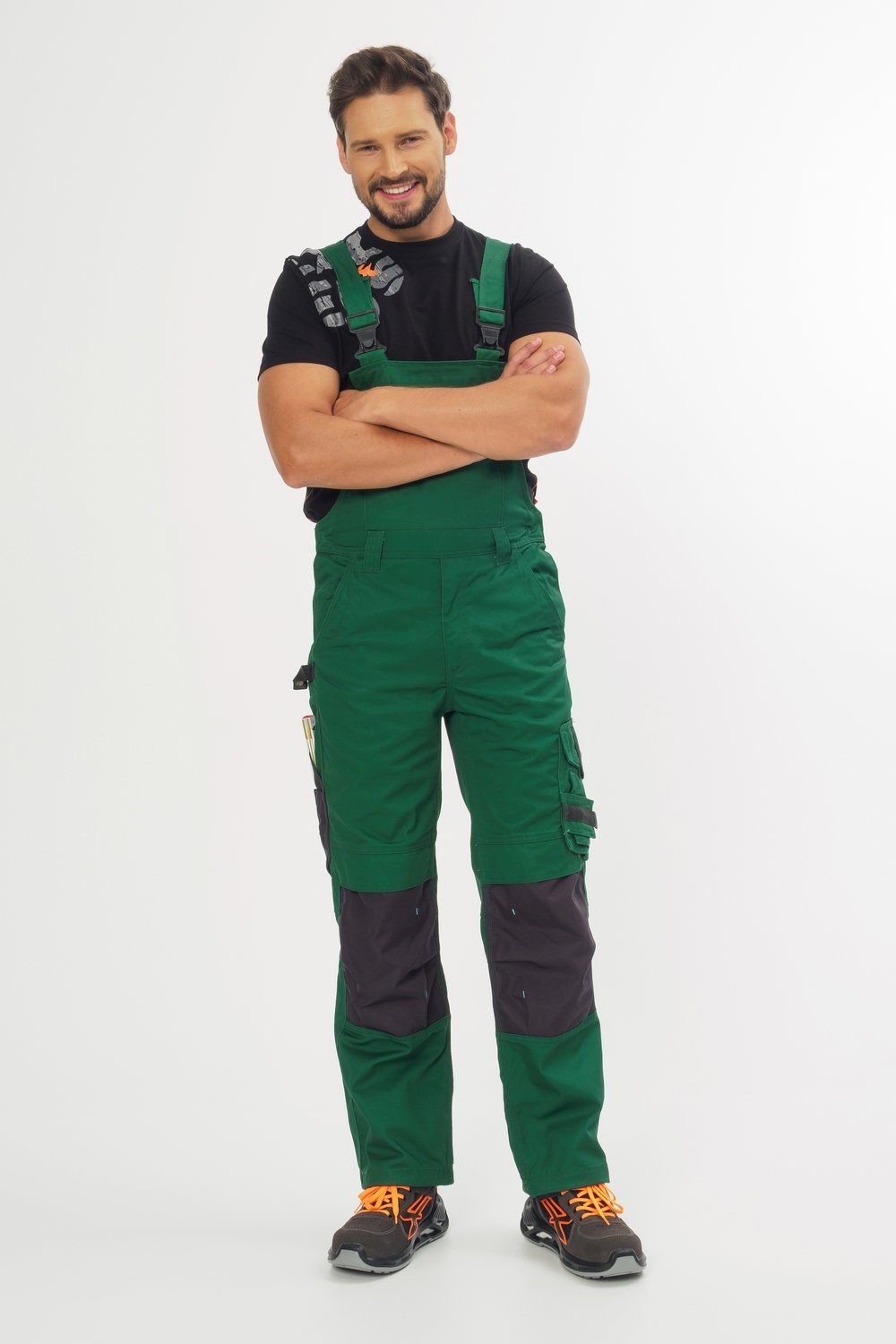 Handwerker in grüner Latzhose mit großer Brusttasche, seitlichen Cargotaschen und verstellbaren Trägern. Dazu ein schwarzes T-Shirt, geeignet für Arbeiten im Handwerk, Bau und Gartenbereich.