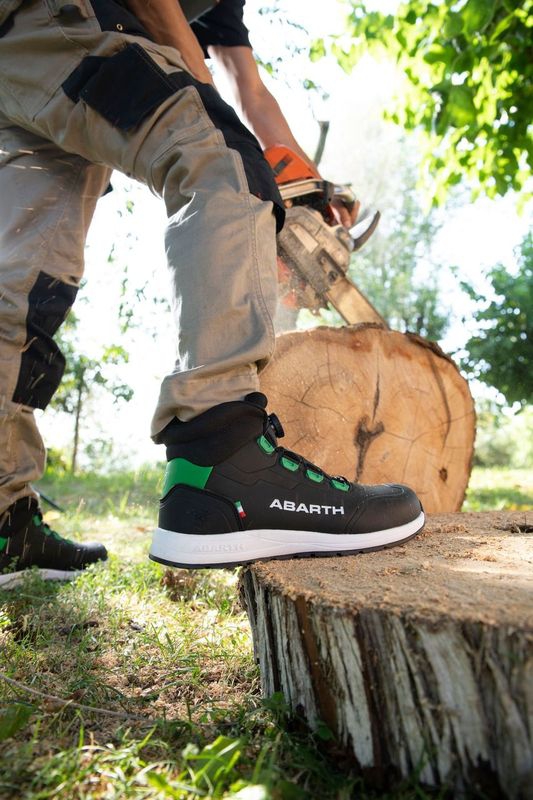 Arbeiter trägt schwarzen ABARTH Sicherheitsschuh mit grünen Details beim Holzsägen auf einem Baumstamm.