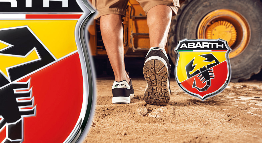 Person in kurzen Hosen geht auf sandigem Boden neben einem gelben Fahrzeug, trägt schwarze Sneaker mit Abarth-Logo. Links und rechts im Bild sind vergrößerte Abarth-Logos eingeblendet.
