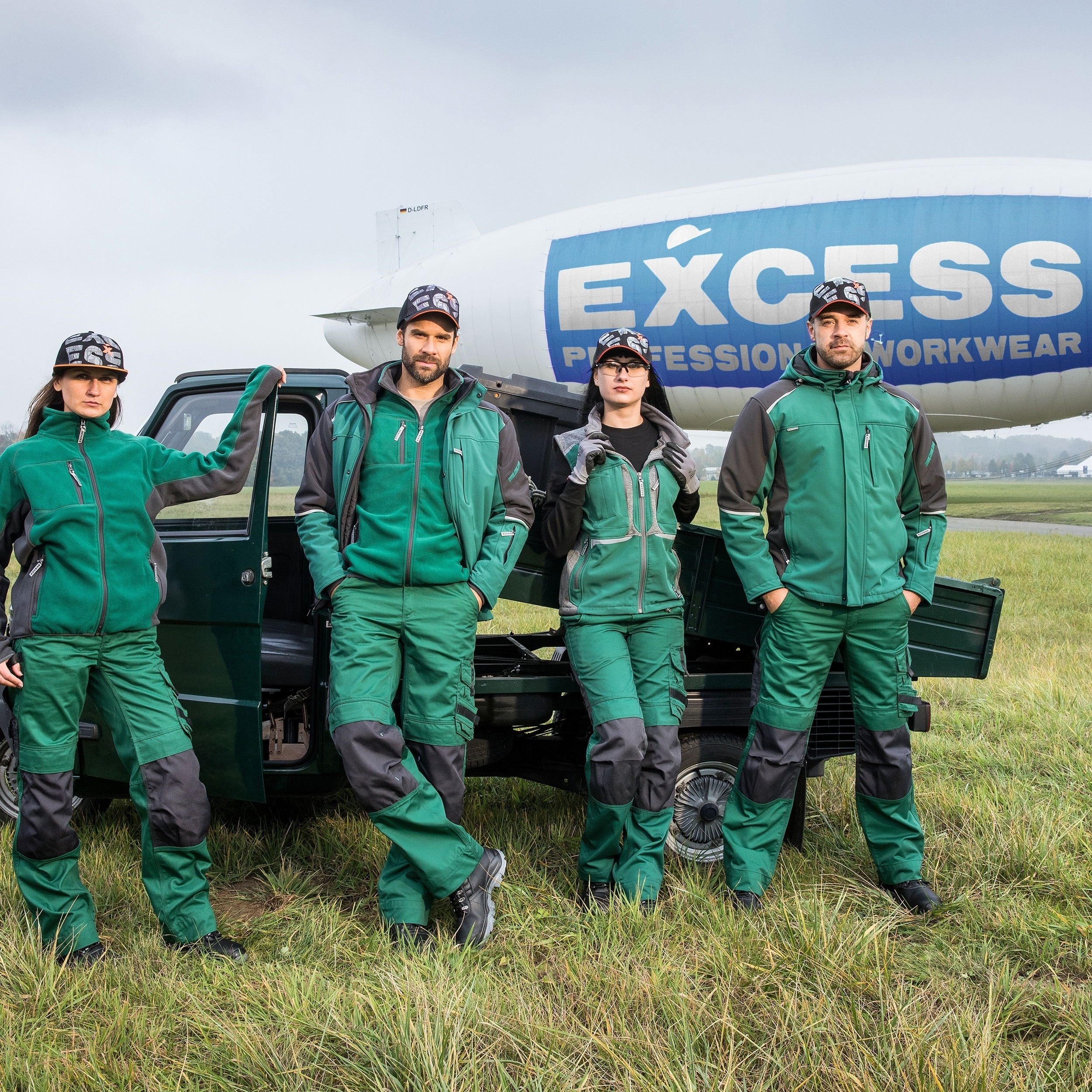 EXCESS Workwear – grüne Arbeitskleidung für Profis, präsentiert von vier Personen auf einer Wiese mit Fahrzeug und Luftschiff.