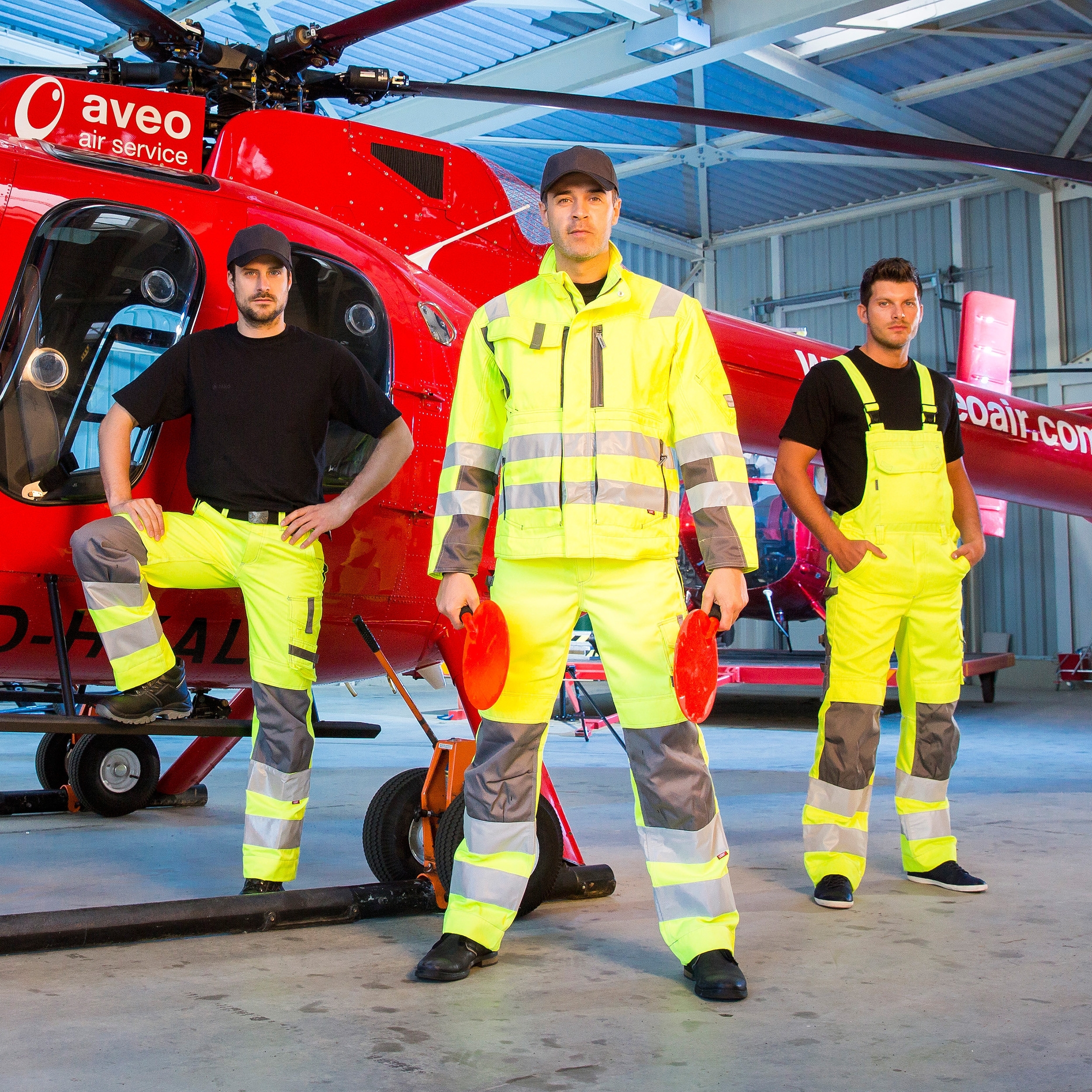 Gruppe von Männern in gelber Arbeits- und Warnschutzkleidung mit Reflexstreifen vor einem roten Hubschrauber im Hangar.
