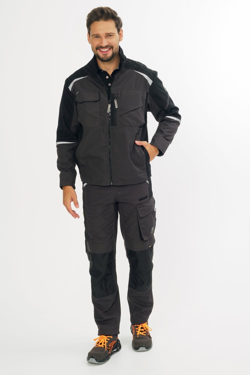 Mann trägt schwarze Arbeitsjacke mit Reißverschlüssen und passende schwarze Arbeitshose mit Taschen und Verstärkungen.