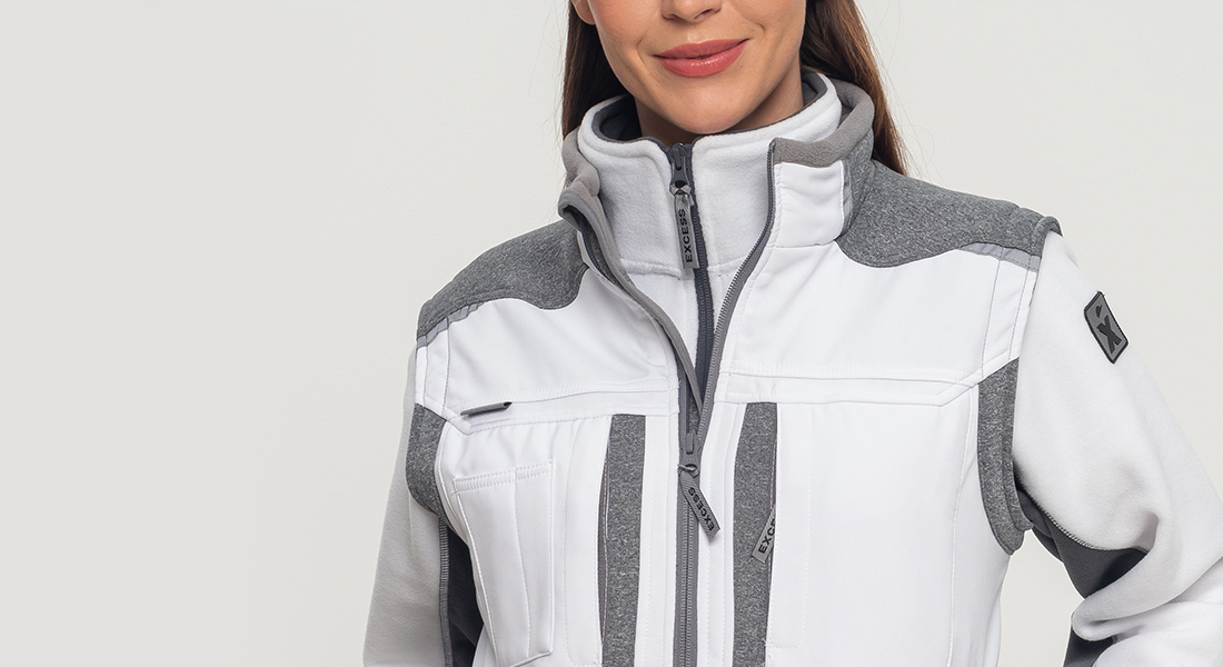 Frau trägt eine weiße und graue Arbeitsjacke mit hohem Kragen und Reißverschlüssen. Der Hintergrund ist neutral hellgrau.