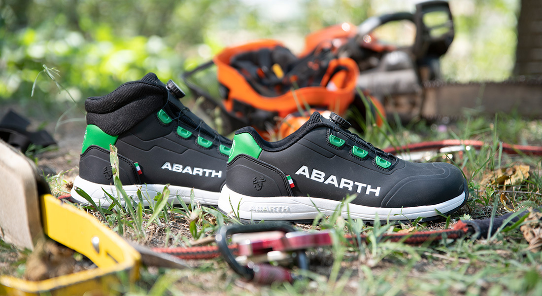 Schwarze Abarth-Arbeitsschuhe mit grünen Details liegen auf einer Wiese zwischen Werkzeugen und einer Motorsäge im Hintergrund.