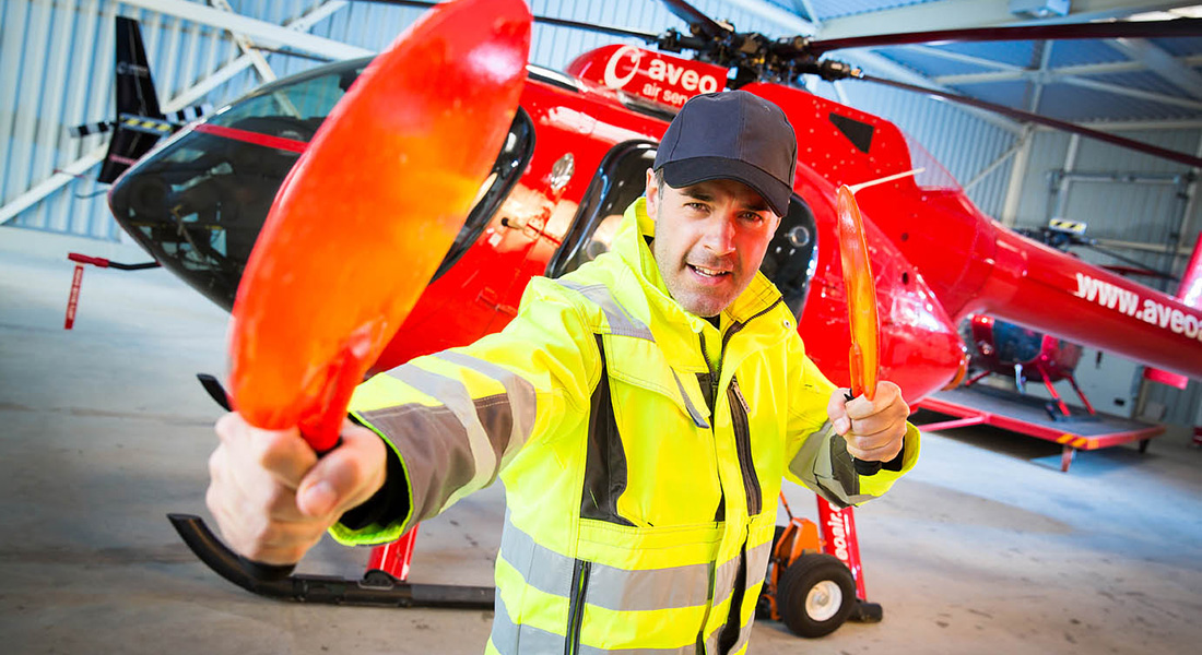 Mann in gelber Warnschutzjacke steht in einer Flughalle vor einem roten Hubschrauber und hält zwei orangefarbene Signalstäbe in den Händen.