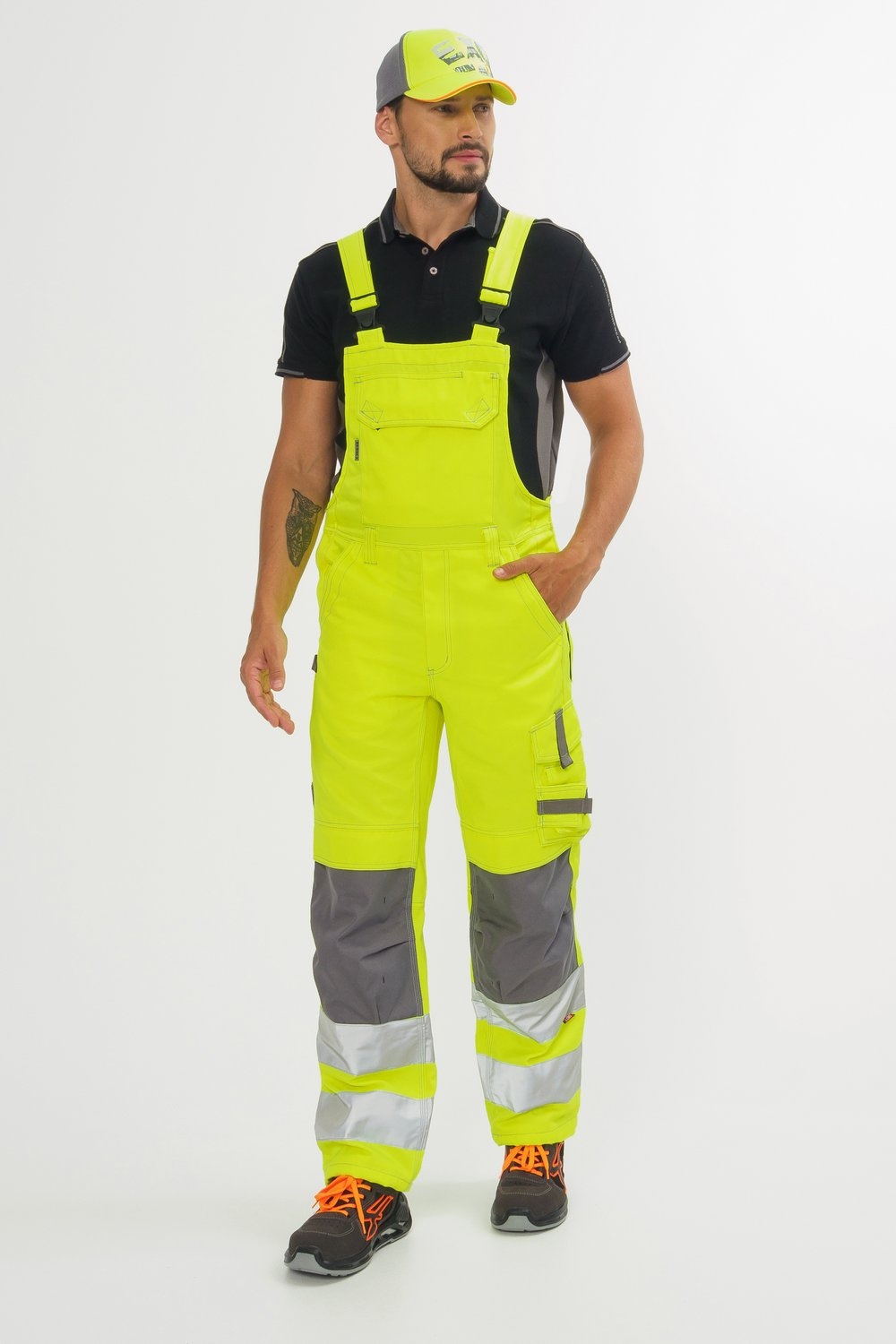 Mann trägt gelbe Latzhose mit silbernen Reflexstreifen, grauen Einsätzen, schwarzem Poloshirt, gelber Kappe und schwarzen Sicherheitsschuhen.