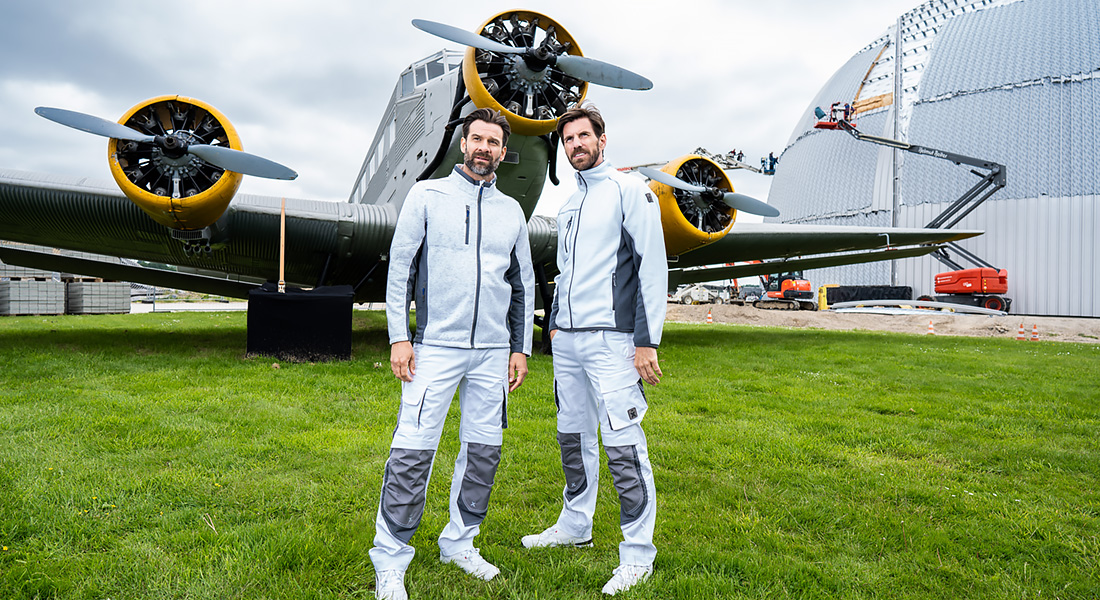 Zwei Männer in weiß-grauer Arbeitskleidung stehen auf einer Wiese vor einem historischen Flugzeug mit gelben Triebwerken und geöffnetem Hangar im Hintergrund.