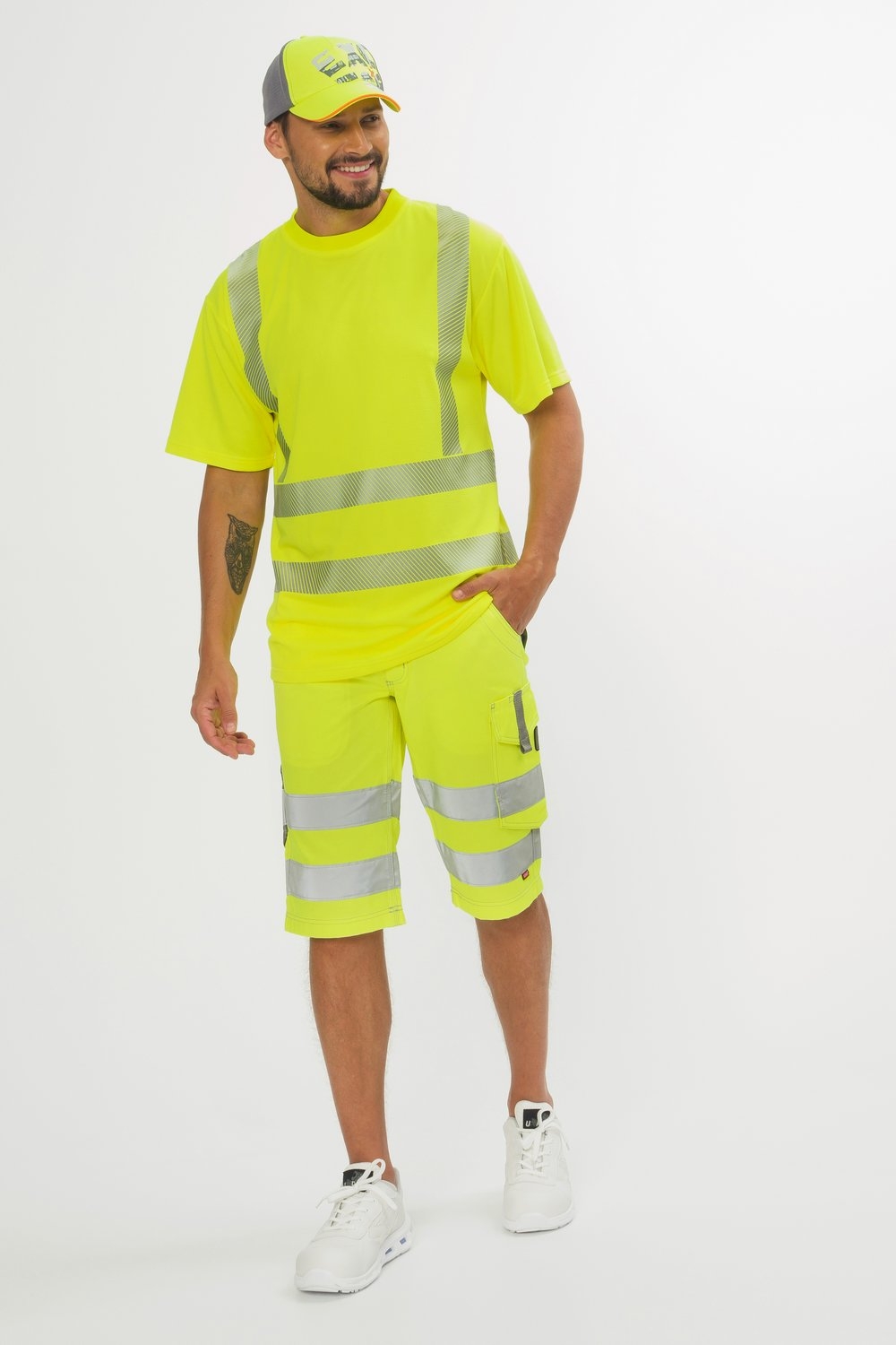 Mann trägt gelbe Warnschutz-Bermudas mit Reflexstreifen, passendes T-Shirt, Kappe und weiße Sicherheitsschuhe.