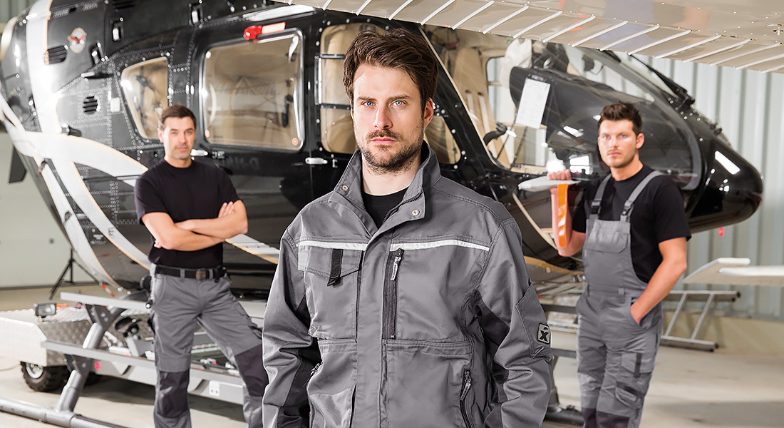 Drei Männer in grauer Arbeitskleidung stehen in einer Werkhalle vor einem schwarzen Hubschrauber. Der vordere Mann blickt direkt in die Kamera.