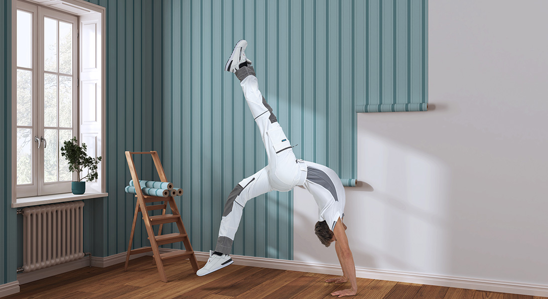 Mann in weiß-grauer Arbeitskleidung steht auf den Händen in einem hellen Raum mit blauer Holzverkleidung, Leiter und Tapetenrolle im Hintergrund.