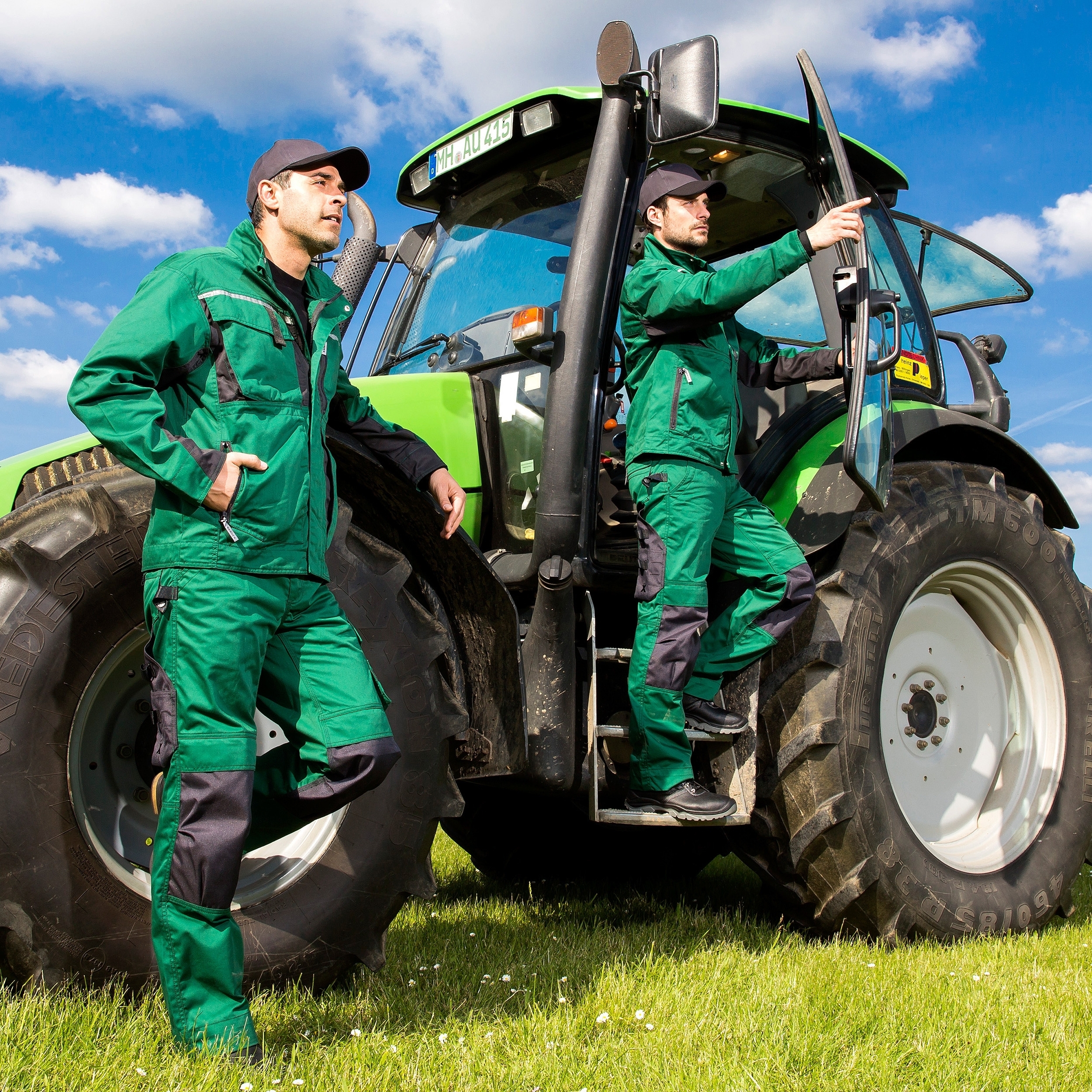 Grüne Arbeitskleidung für Landwirtschaft – zwei Personen mit Berufsmode von EXCESS vor modernem Traktor auf Feldwiese.