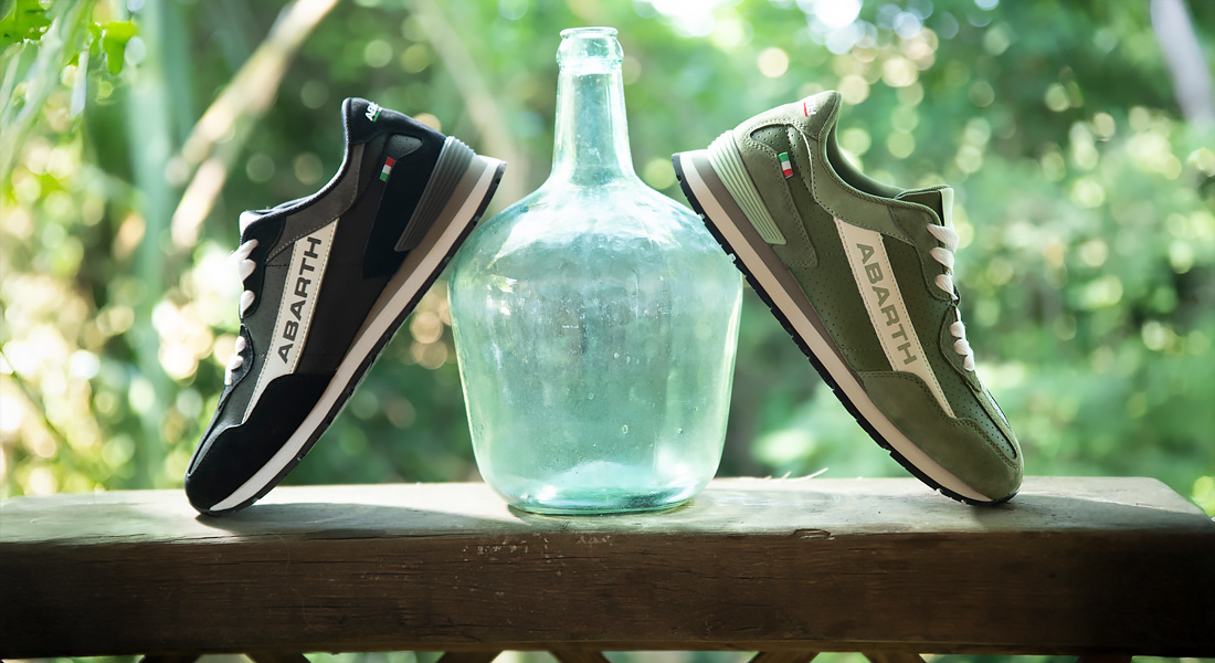 Zwei Sneaker der Marke Abarth, ein schwarzer und ein olivgrüner, stehen auf einem Holzgeländer beidseitig einer großen Glasflasche. Im Hintergrund ist unscharf ein grüner, sonnendurchfluteter Garten zu sehen.