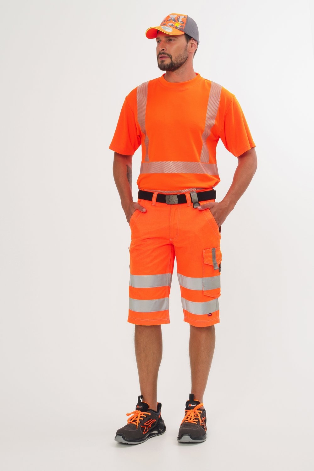 Mann trägt orange Warnschutz-Bermudas mit Reflexstreifen, passendes T-Shirt, Kappe und schwarze Sicherheitsschuhe mit orangen Details.